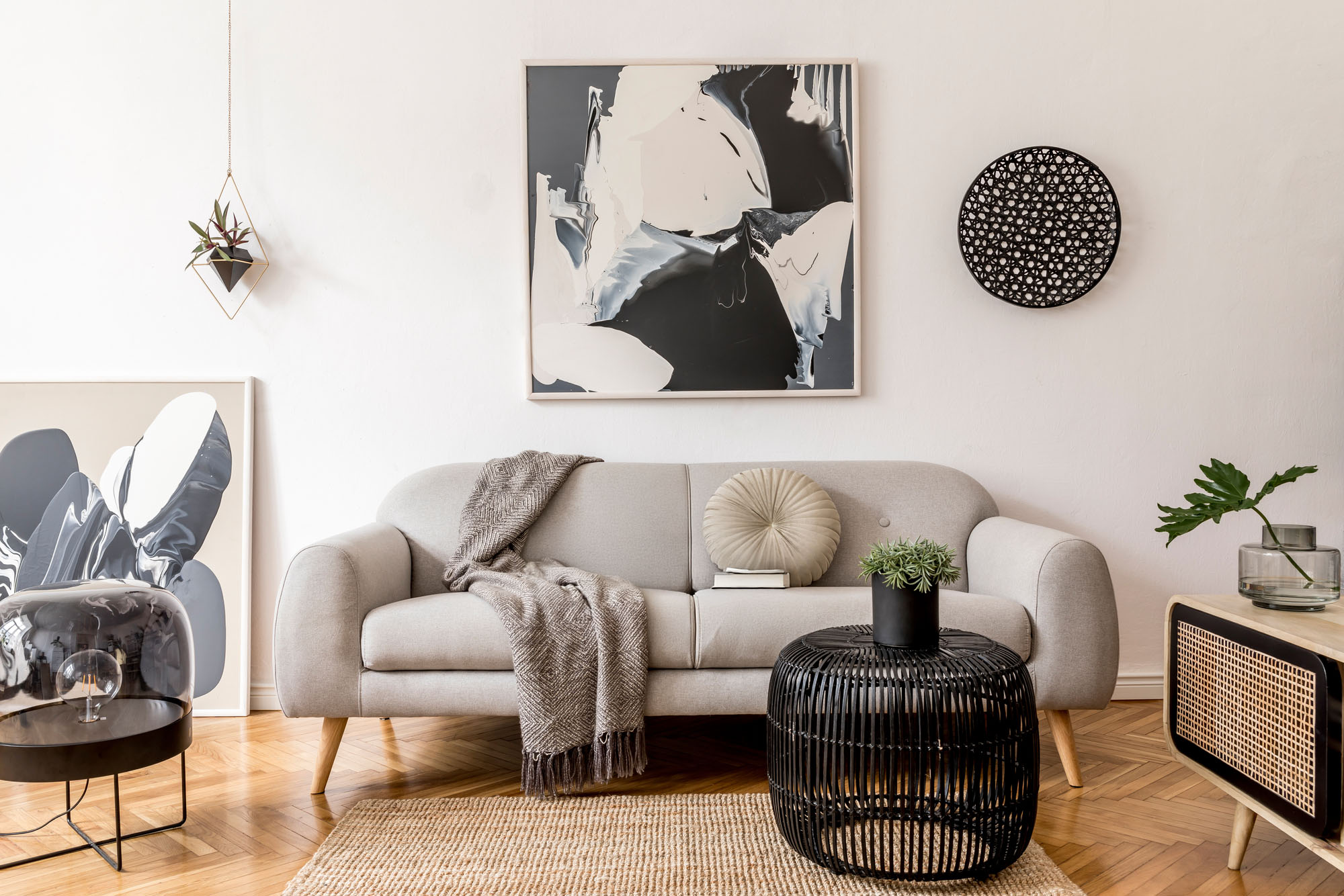 Stylish and scandinavian living room interior of modern apartment with gray sofa, design wooden commode, black table, lamp, abstract paintings on the wall. Beautiful dog lying on the couch. Home decor.